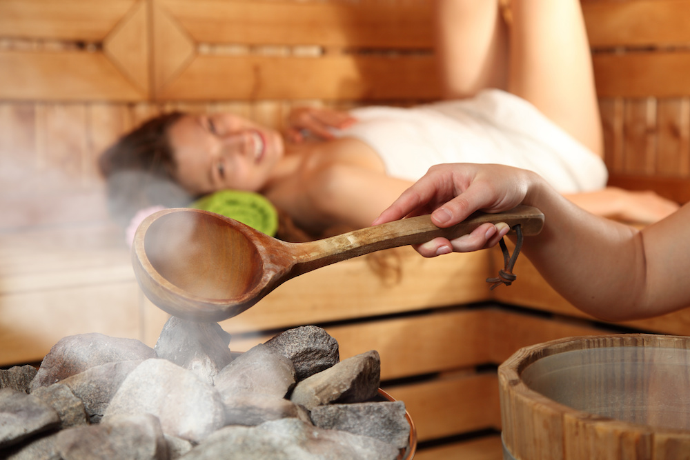 Woman laying in sauna with hand pouring water on sauna stones creating steam at spa
